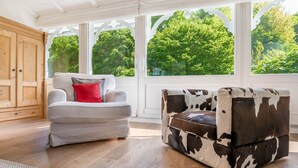 Um quarto com um guarda-roupa de madeira, um sofá cinza com uma almofada vermelha e um banquinho com padrão de couro de vaca.