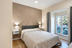 Ein Schlafzimmer mit Bett, Nachttischlampen, einem Nachttisch, einem Fenster mit Blick auf die Straße und einem Balkon.