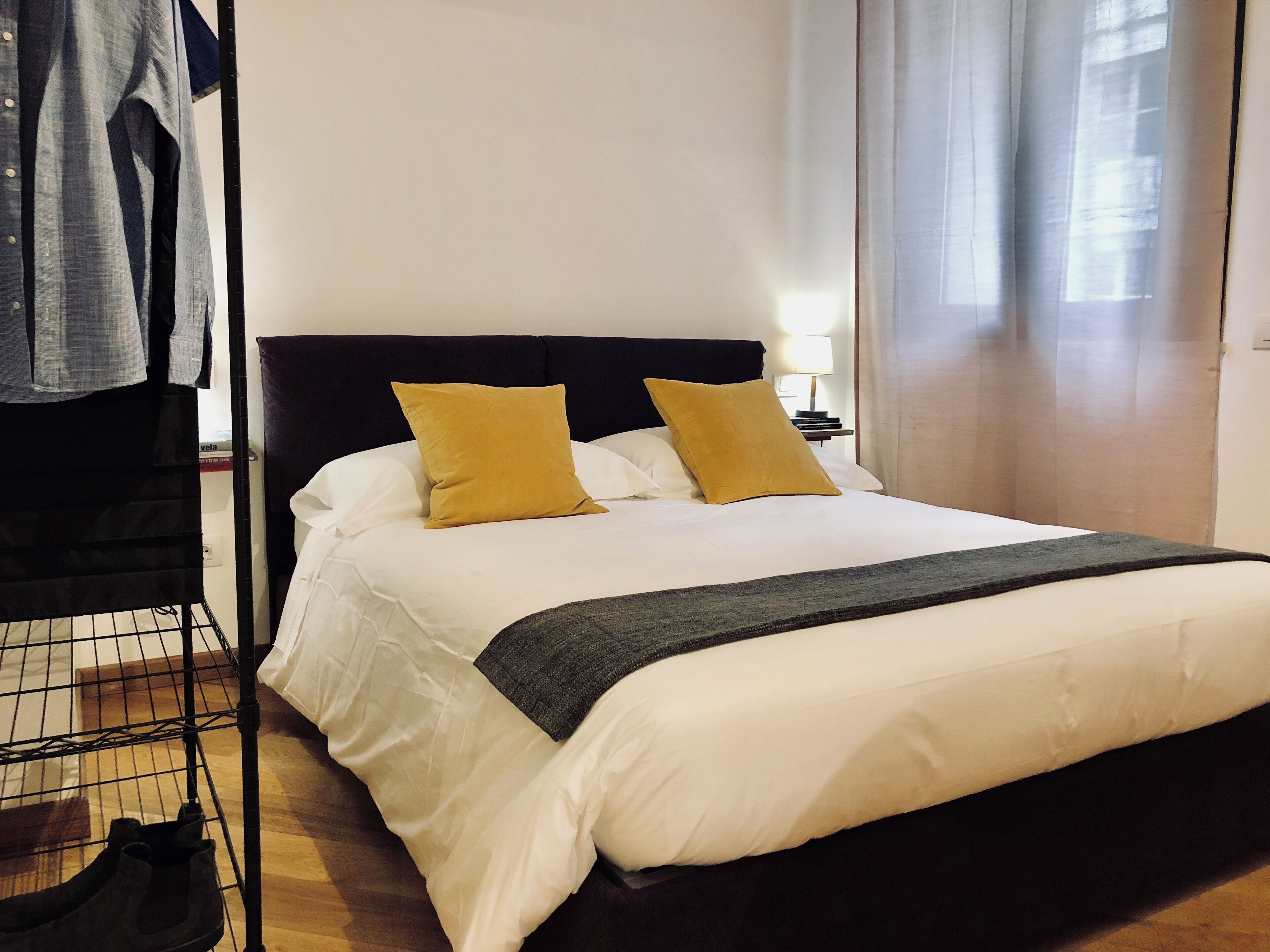Ein ordentlich bezogenes Bett mit weißer Bettwäsche und zwei senfgelben Kissen, ein schwarzer Kopfteil und eine graue Tagesdecke.