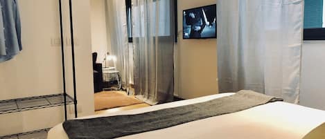 A hotel room with a bed, a TV mounted on the wall, a wardrobe, and a small table with a lamp.