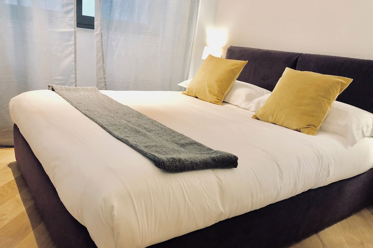 A neatly made bed with white and yellow pillows, a dark grey throw blanket, and a wooden floor.