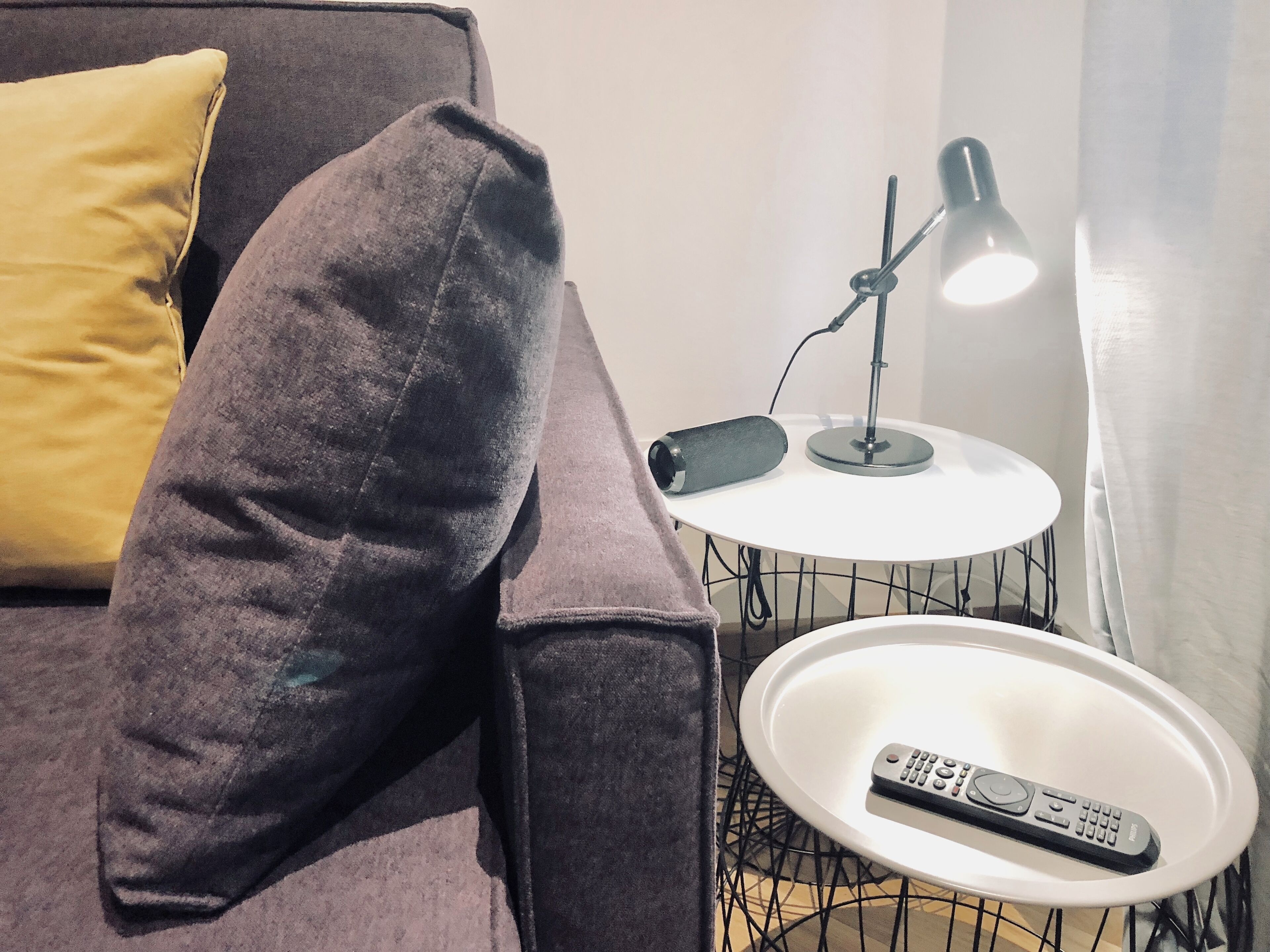 A dark gray sofa with a yellow pillow, a white round side table with a black lamp, and a remote control on the table.