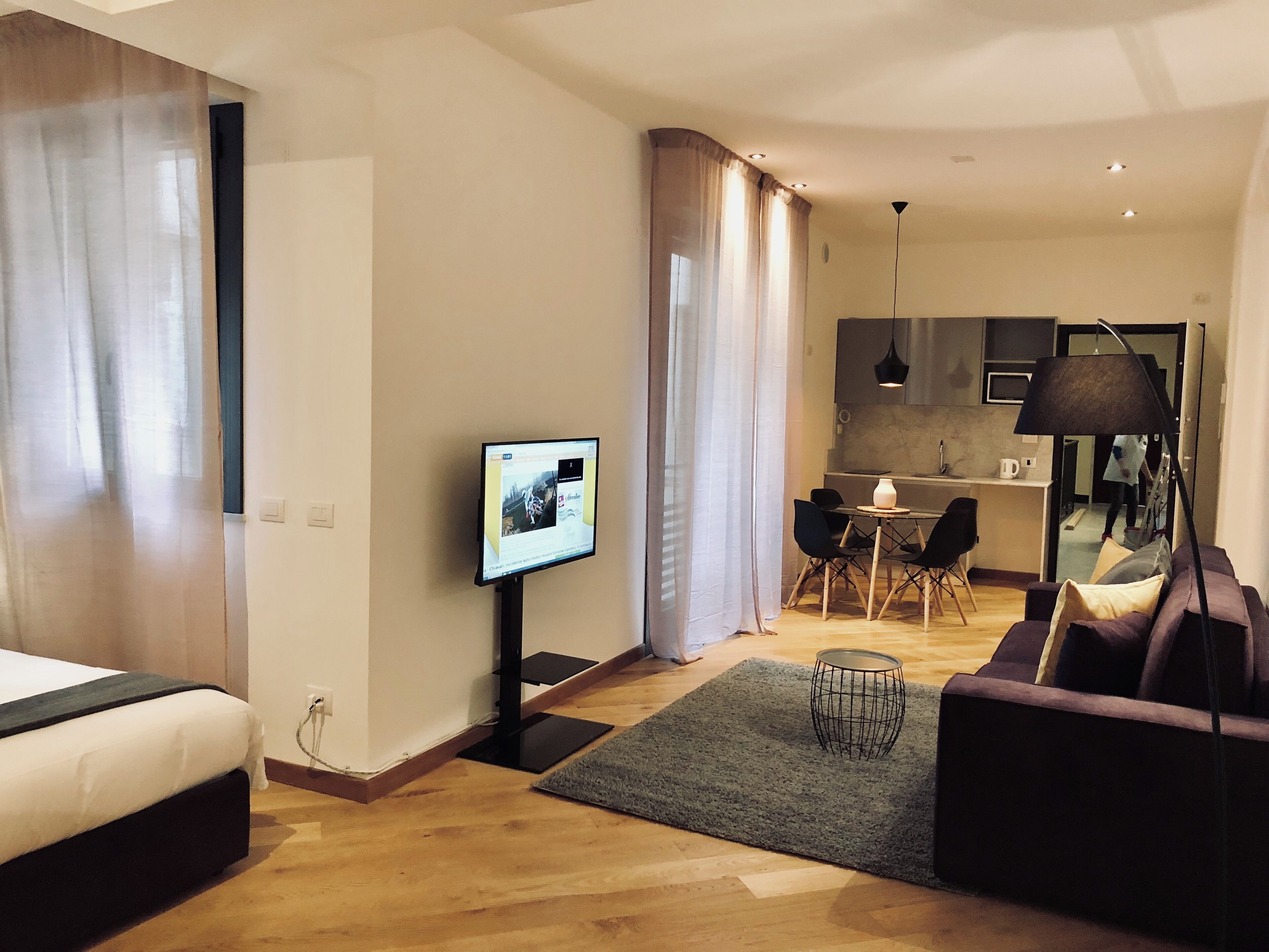 A modern living room with a sofa, a TV mounted on the wall, a dining area with chairs, and a kitchen in the background.