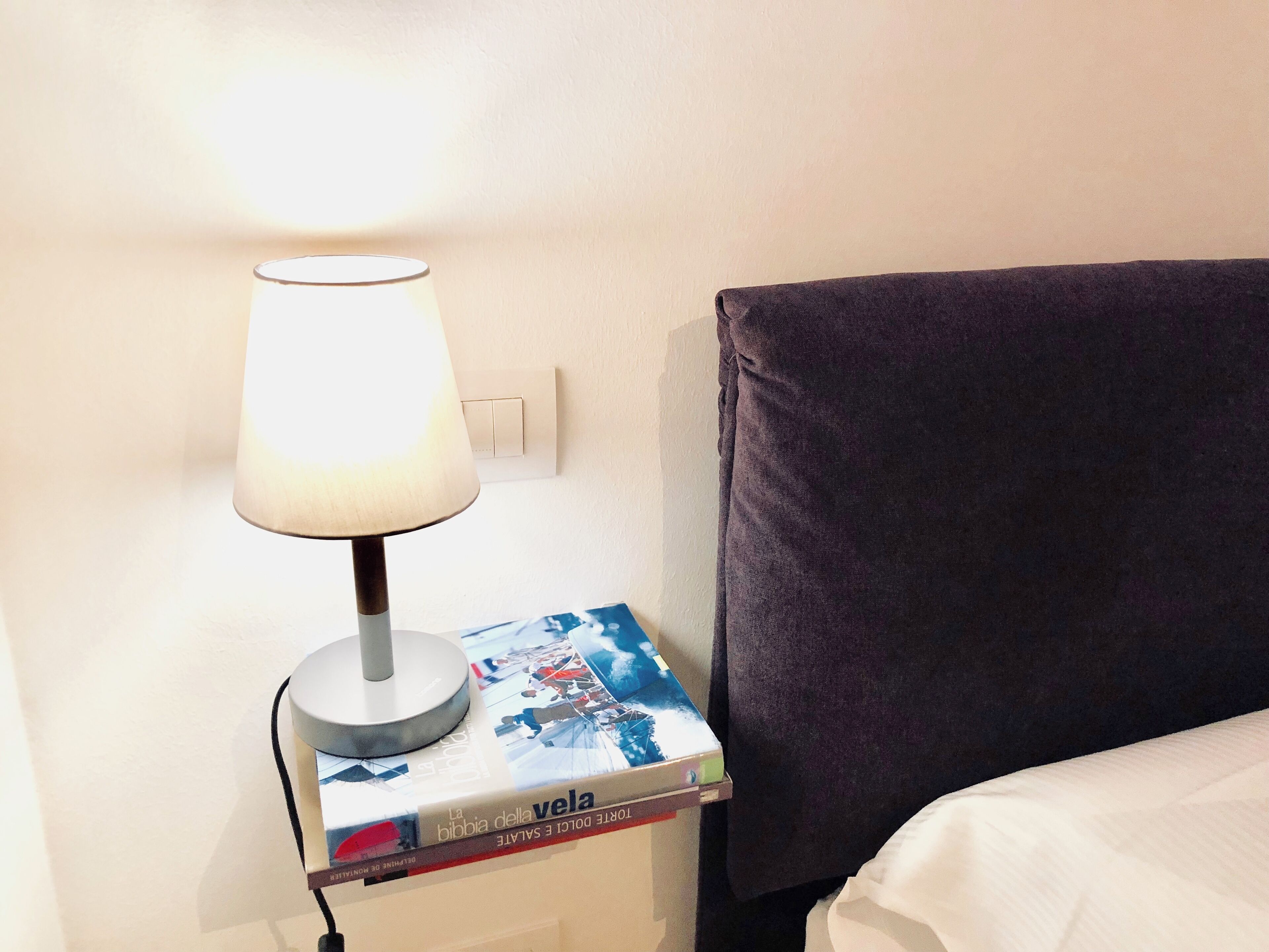 A bedside table with a lamp, a book titled 'La bibbia della vela', and a dark-colored pillow on a bed.