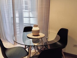 Una mesa de vidrio con un mantel tejido, rodeada de sillas negras, en una habitación con ventana y cortinas.