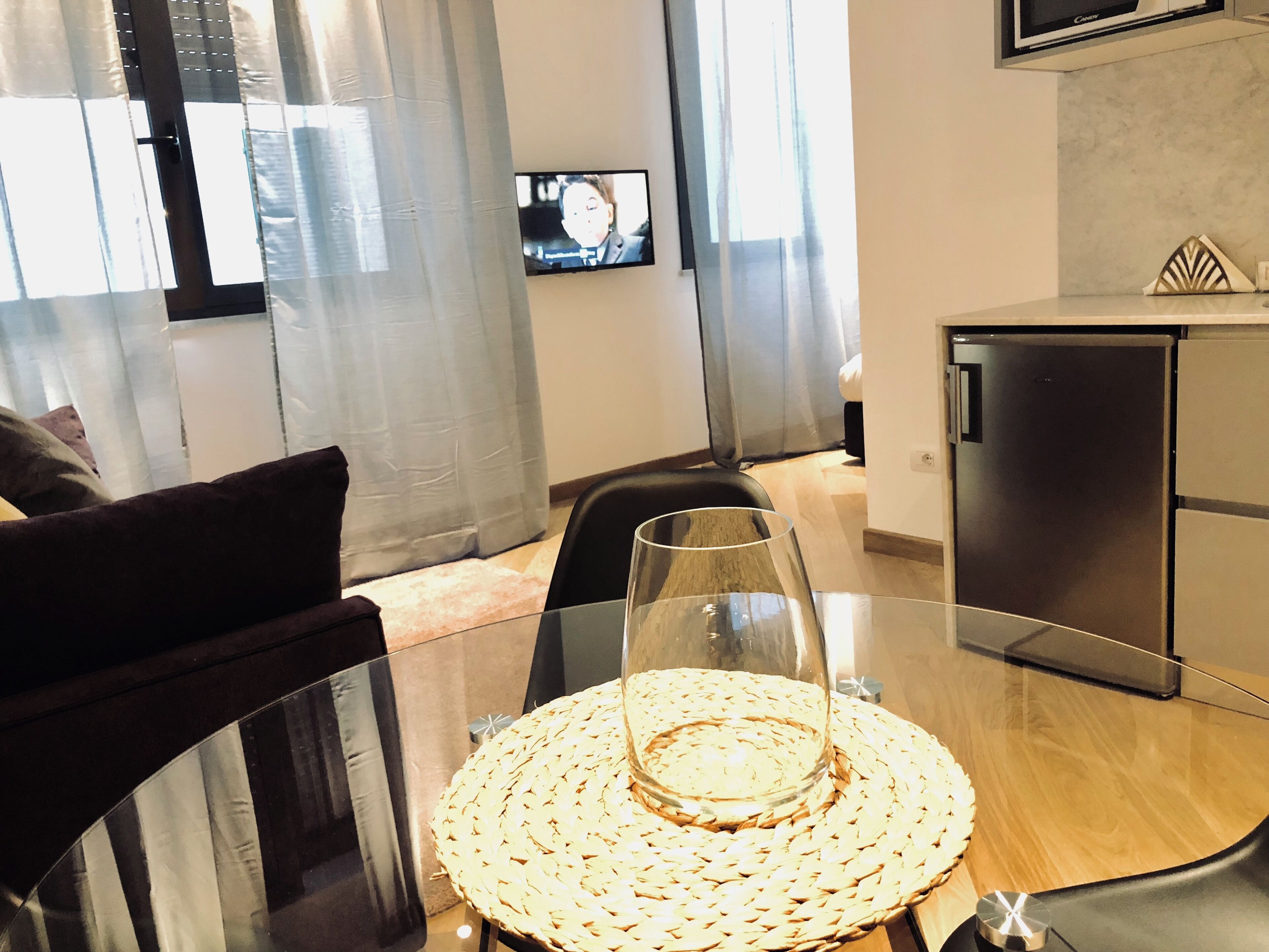 Ein modernes Wohnzimmer mit einem Glastisch, einer gewebten Tischmatte, einem schwarzen Sofa, einem an der Wand befestigten Fernseher und einem Kühlschrank.