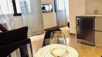 Ein modernes Wohnzimmer mit einem Glastisch, einer gewebten Tischmatte, einem schwarzen Sofa, einem an der Wand befestigten Fernseher und einem Kühlschrank.