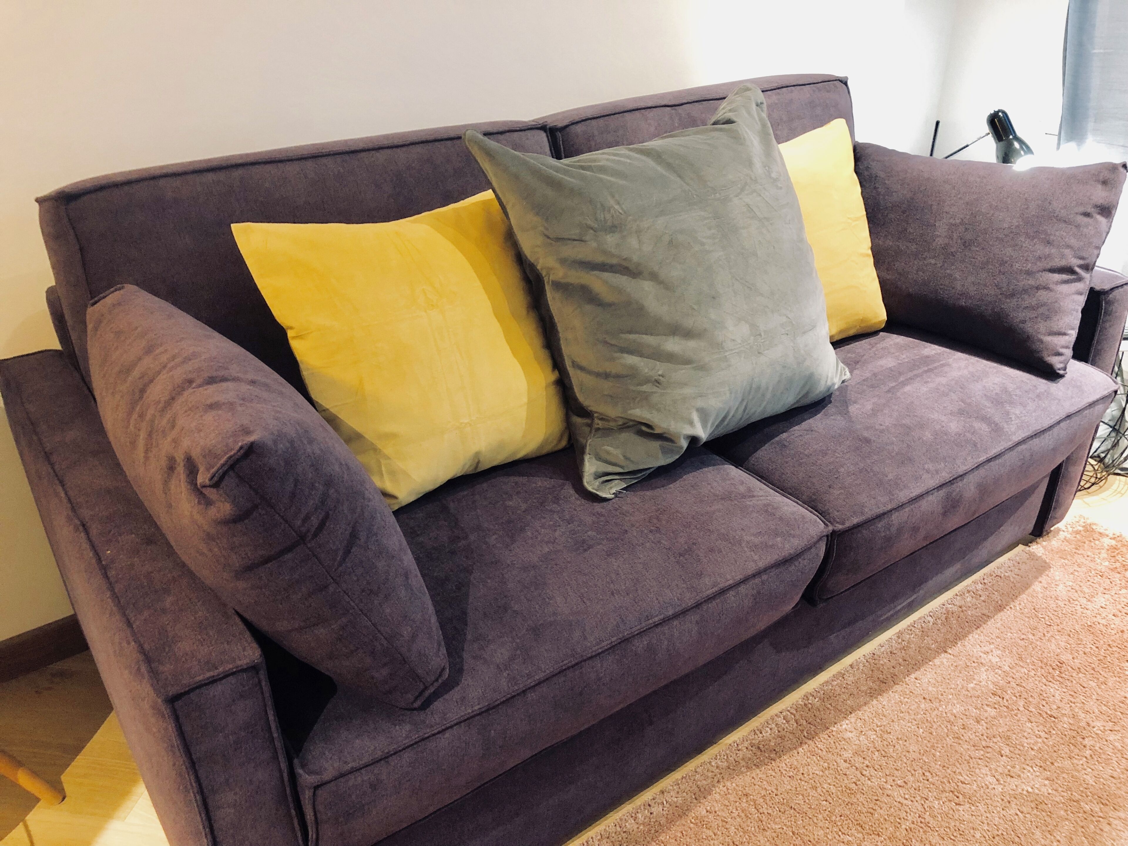A purple sofa with two pillows, one yellow and one gray, in a room with a wooden floor and a window.
