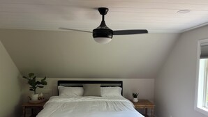 A bedroom with a bed, a ceiling fan, a nightstand with a plant, and a window.