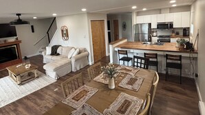 A modern kitchen with a central island, dining area with a wooden table and chairs, and a living room with a sofa and fireplace.