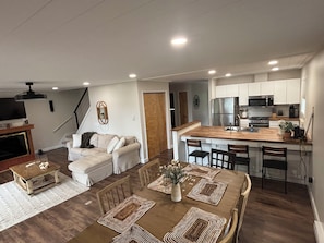 A modern kitchen with a central island, dining area with a wooden table and chairs, and a living room with a sofa and fireplace.