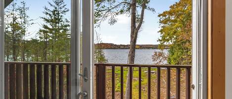 A view from a sliding glass door to a deck with a wooden railing overlooking a lake and trees.