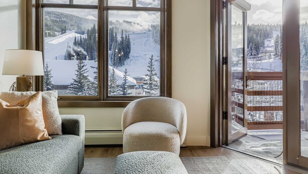 Une pièce chaleureuse avec une grande fenêtre donnant sur un paysage montagneux enneigé, un fauteuil confortable et un canapé agrémenté de coussins.
