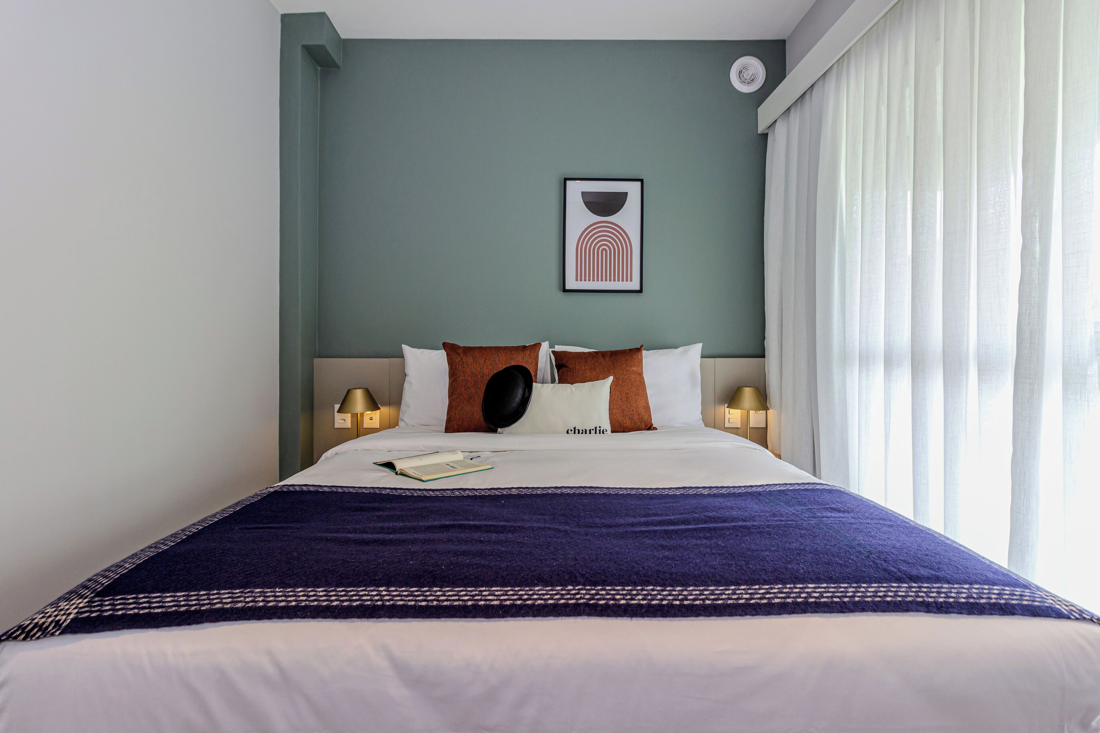 A neatly made bed with a blue blanket and pillows, a framed picture on the wall, and a window with curtains.