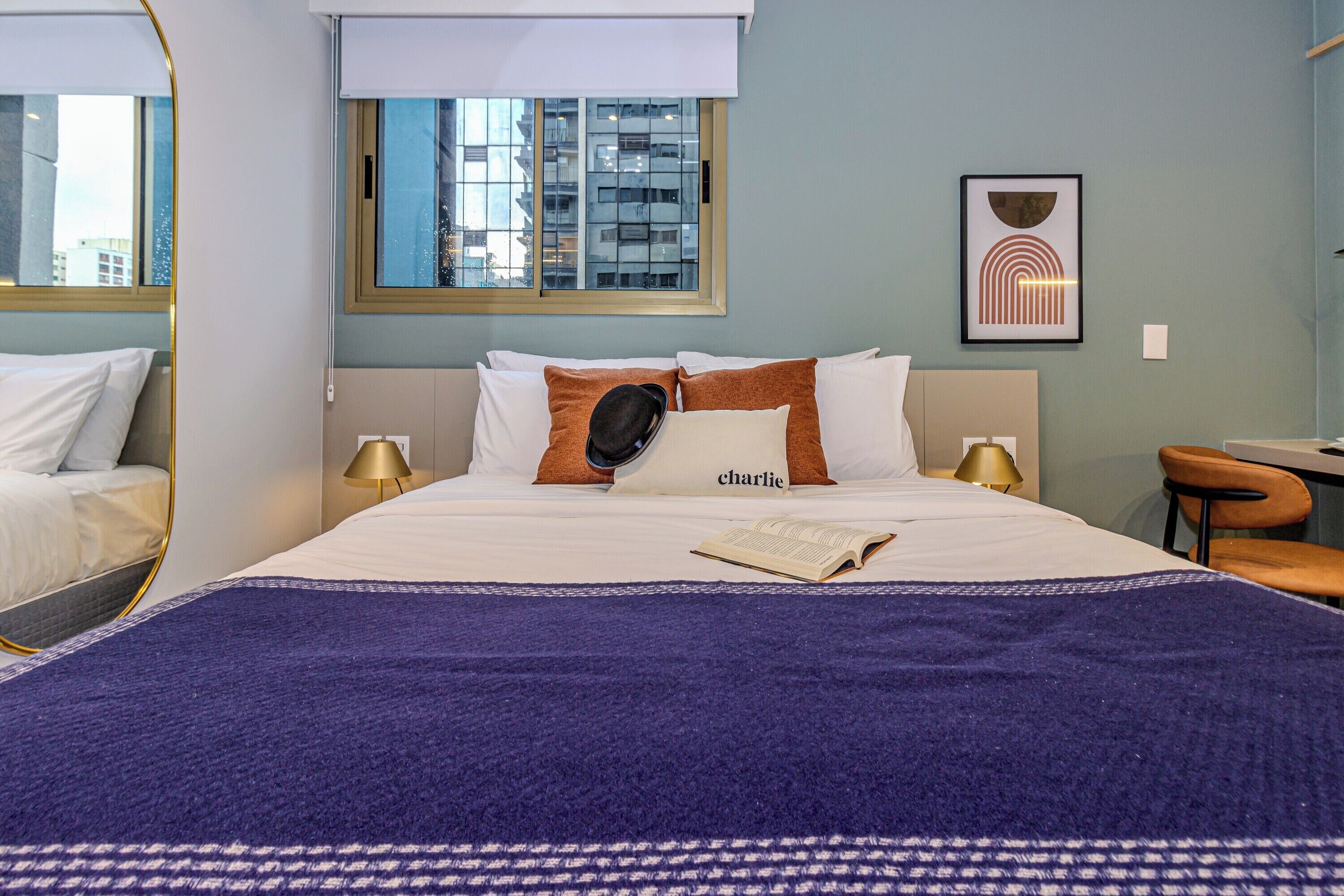 Ein Hotelzimmer mit einem großen Bett, einem Schreibtisch und einem Stuhl. Ein Fenster bietet Blick auf Gebäude, und an der Wand hängt ein gerahmtes Bild.