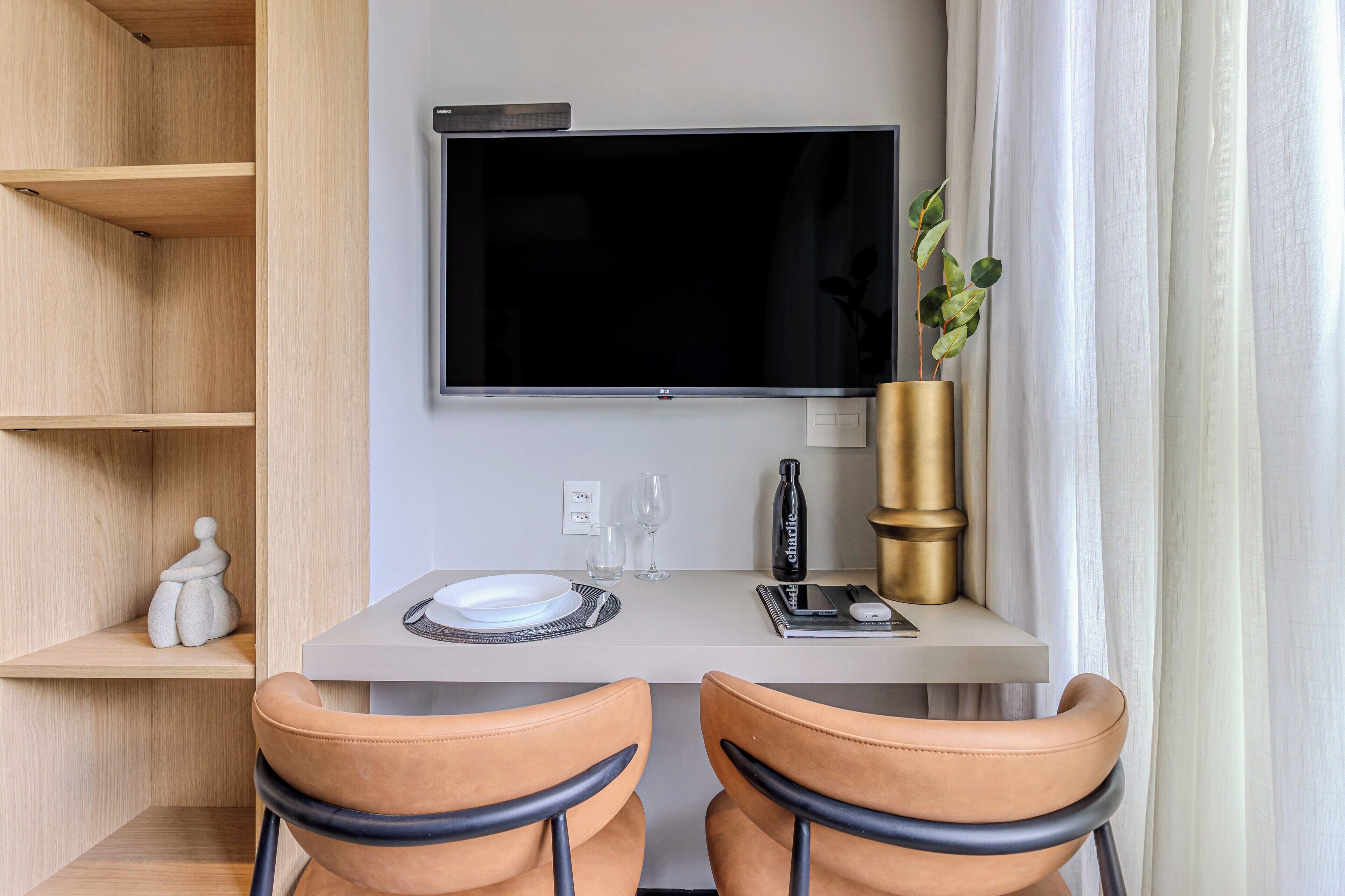 A modern room with a flat-screen TV mounted on the wall, a small table with a plate, glasses, and a notebook, two leather chairs, and a potted plant.