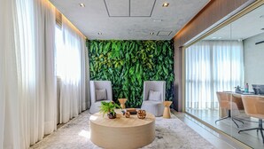 A modern meeting room with a round wooden table, two grey chairs, a green leafy wall, and large windows with white curtains. - Charlie Nik Paradiso \ Soft Opening (São Paulo)