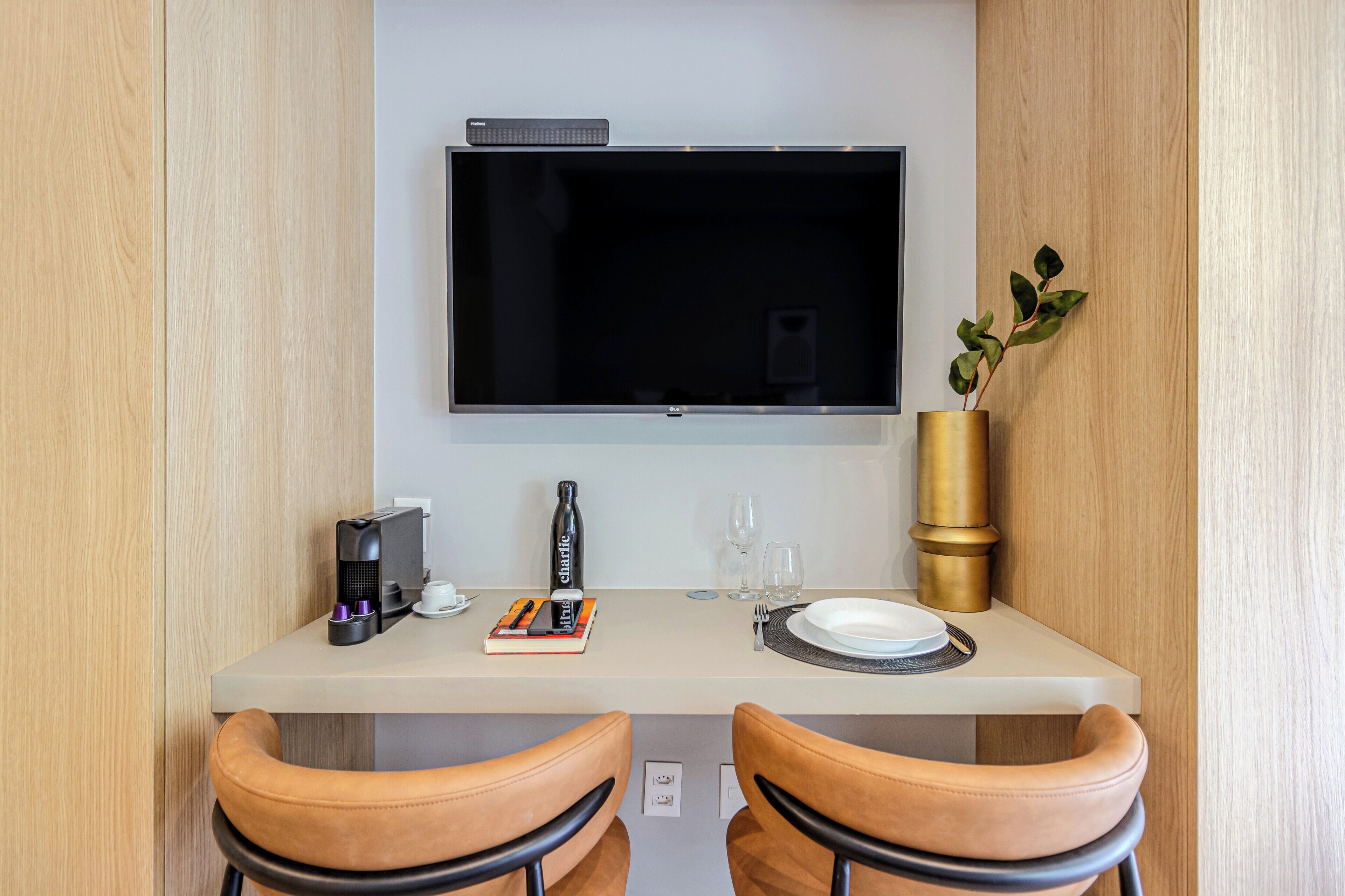 A modern hotel room with a flat-screen TV mounted on the wall, a coffee machine, a book, a bottle of water, and a vase with a plant.