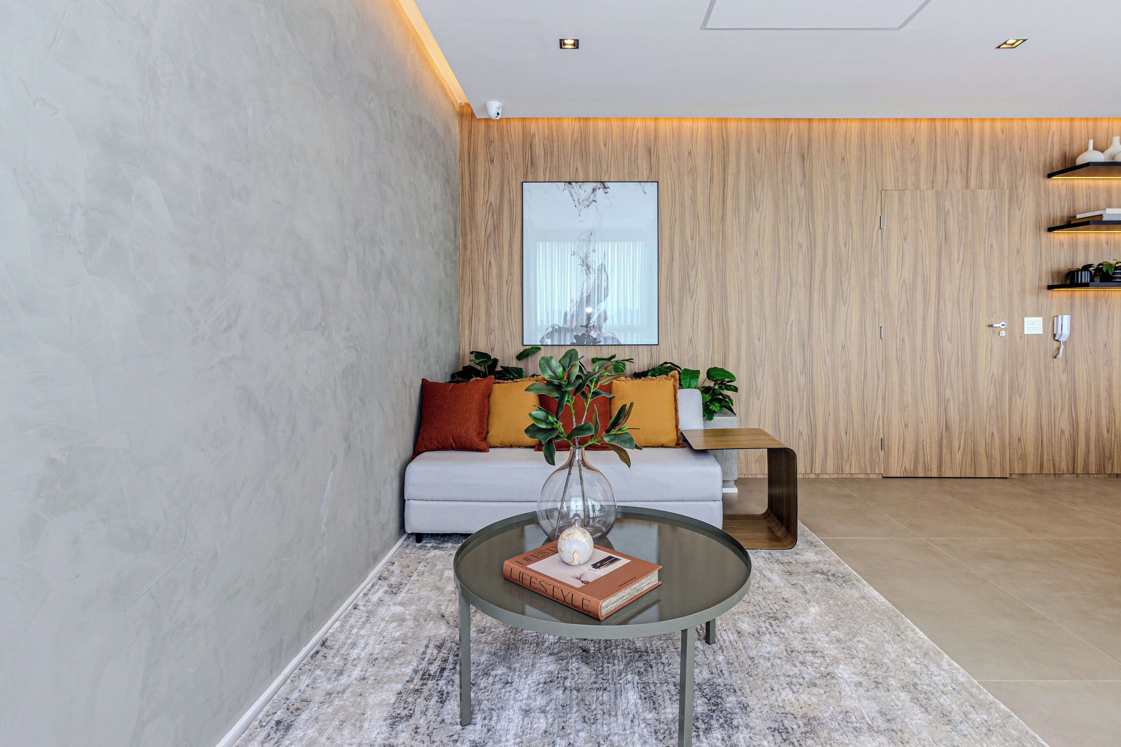 A modern living room with a glass coffee table, a sofa with cushions, a wooden accent wall, and a large window.