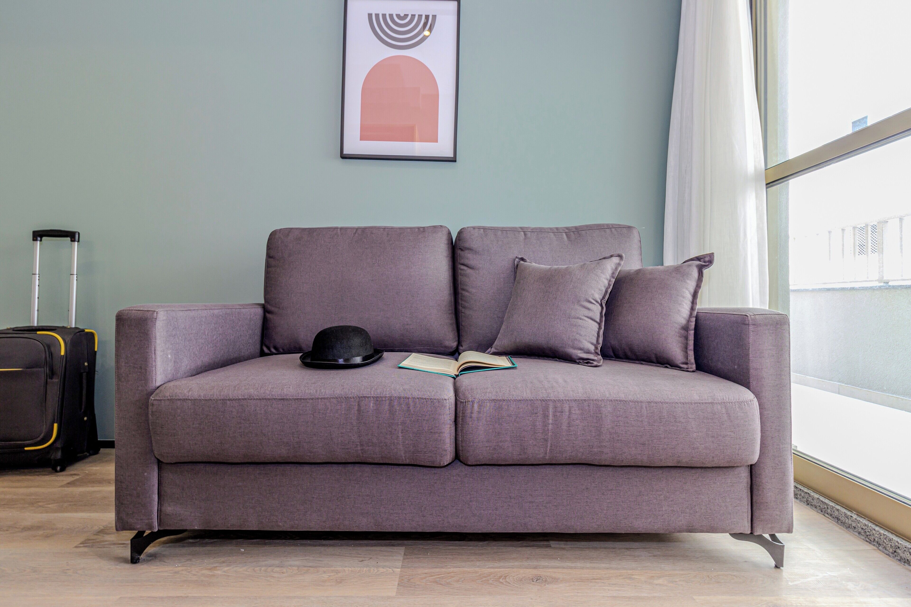 A grey sofa with a black hat and an open book, a suitcase, and a framed picture on the wall.