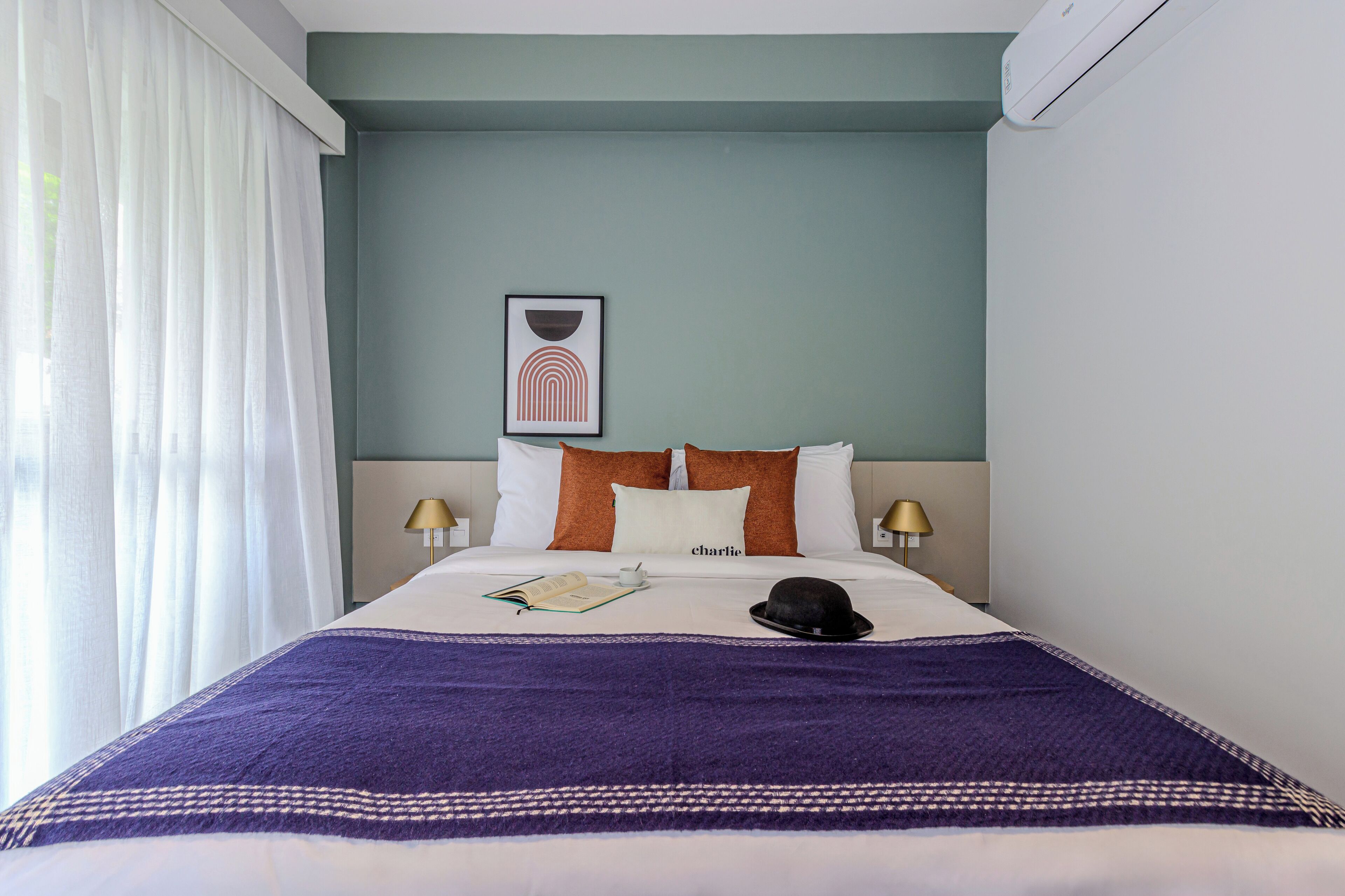 Un letto rifatto con cura, con una coperta viola, un libro e un cappello. Due abat-jour e un quadro incorniciato sulla parete.
