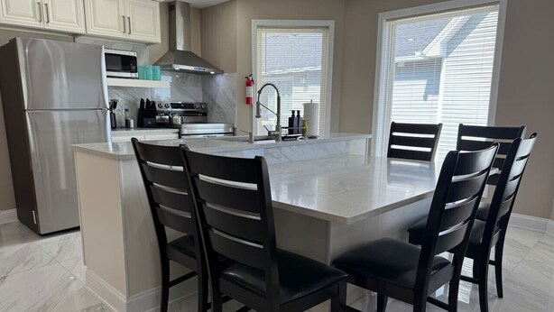 A modern kitchen with a large island, dark chairs, and a stainless steel refrigerator.