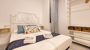 Ein ordentlich bezogenes Bett mit einem weißen Kopfteil, einem blauen Kissen und einem farbenfrohen Blumenkissen. Auf dem Bett liegen zwei weiße Handtücher gefaltet und eine graue Decke. Eine weiße Kommode mit Korbwäsche befindet sich rechts.