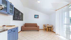 Uma cozinha com armário azul, um quadro-negro com desenhos, uma área de refeições com mesa e cadeiras e uma janela grande com cortinas leves.