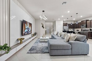 A modern living room with a grey sectional sofa, a flat-screen TV mounted on the wall, and a kitchen area with dark wooden cabinets and stainless steel appliances. - 8BR house sleeps 20 with game room (Clermont)