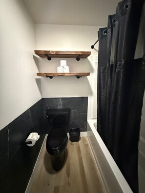 A compact bathroom with a black toilet, a wooden shelf above it, and a black bathtub. - CB&M Downtown Suites (Roosevelt)