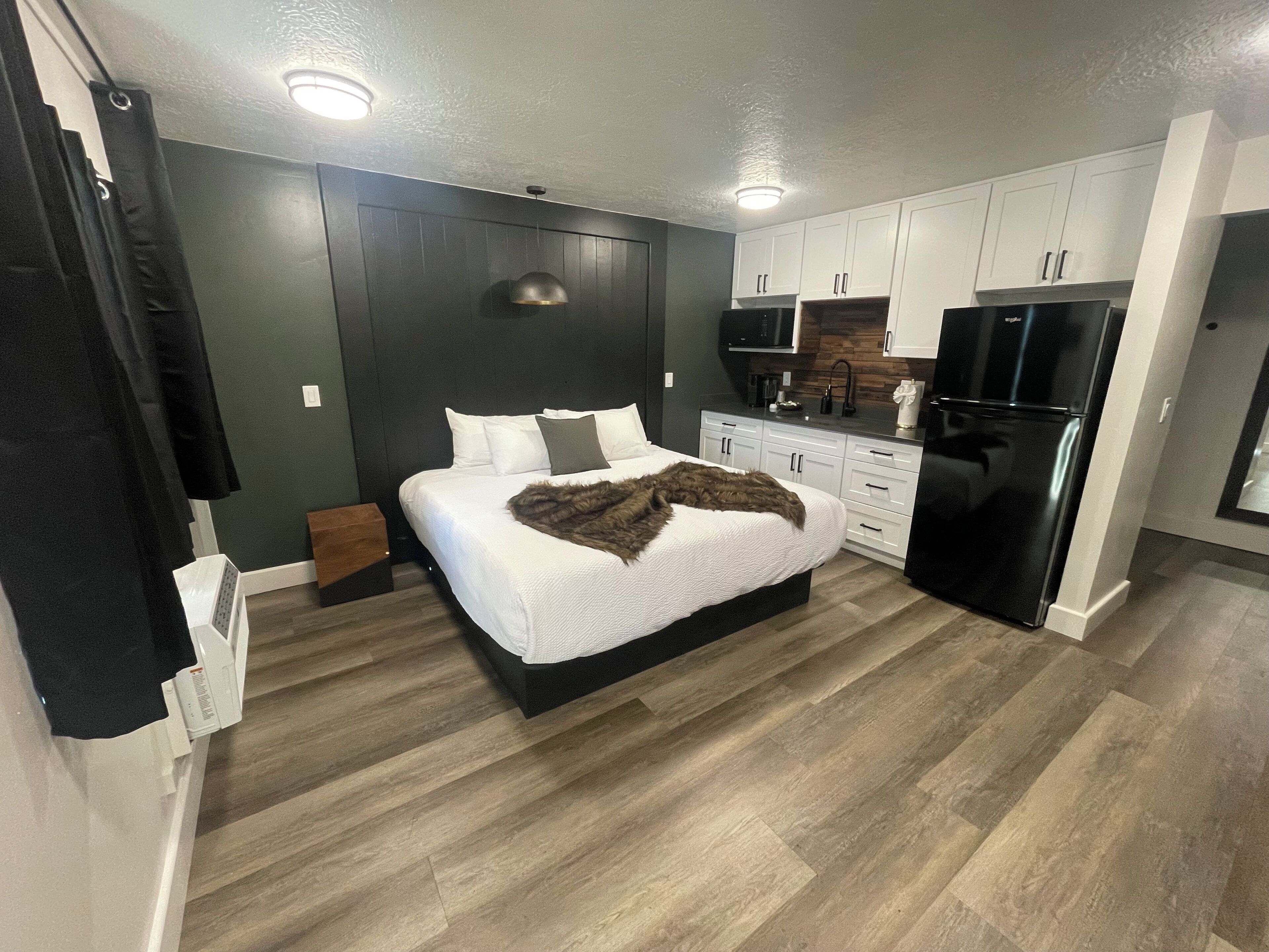 A compact bedroom with a bed, bedside table, and a small seating area. The room features a kitchenette with a refrigerator, microwave, and stove.