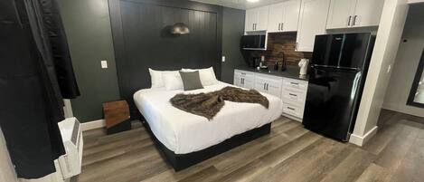 A compact bedroom with a bed, bedside table, and a small seating area. The room features a kitchenette with a refrigerator, microwave, and stove.