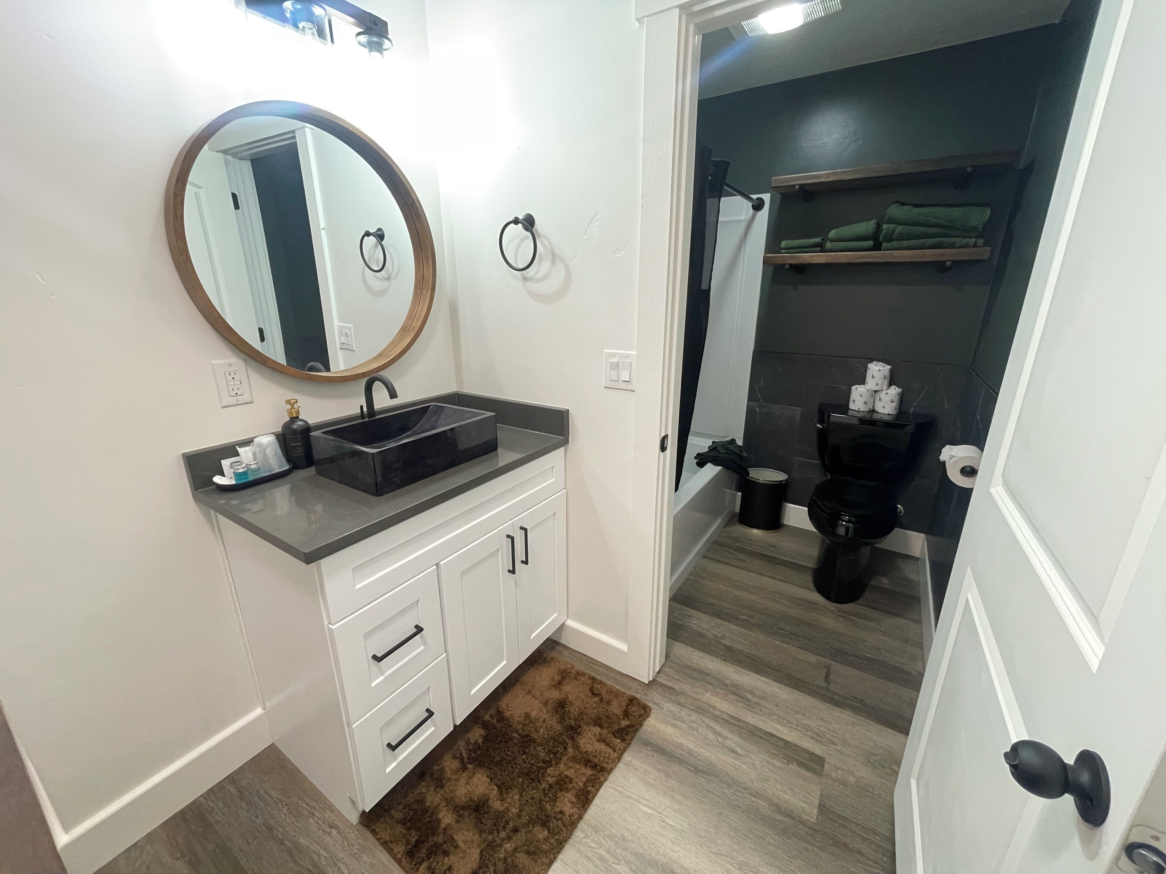 A modern bathroom with a black sink, white cabinets, a round mirror, and a shower area visible through an open door.