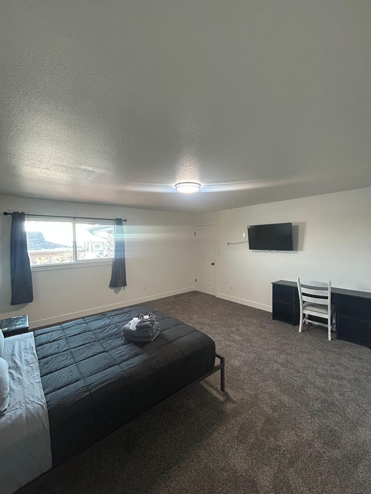 A bedroom with a bed, a desk, a chair, a TV, and a window with curtains.