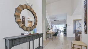 Um corredor com um aparador, um espelho e um banco. Há uma escada com corrimão e uma porta com a placa 'WELCOME'.