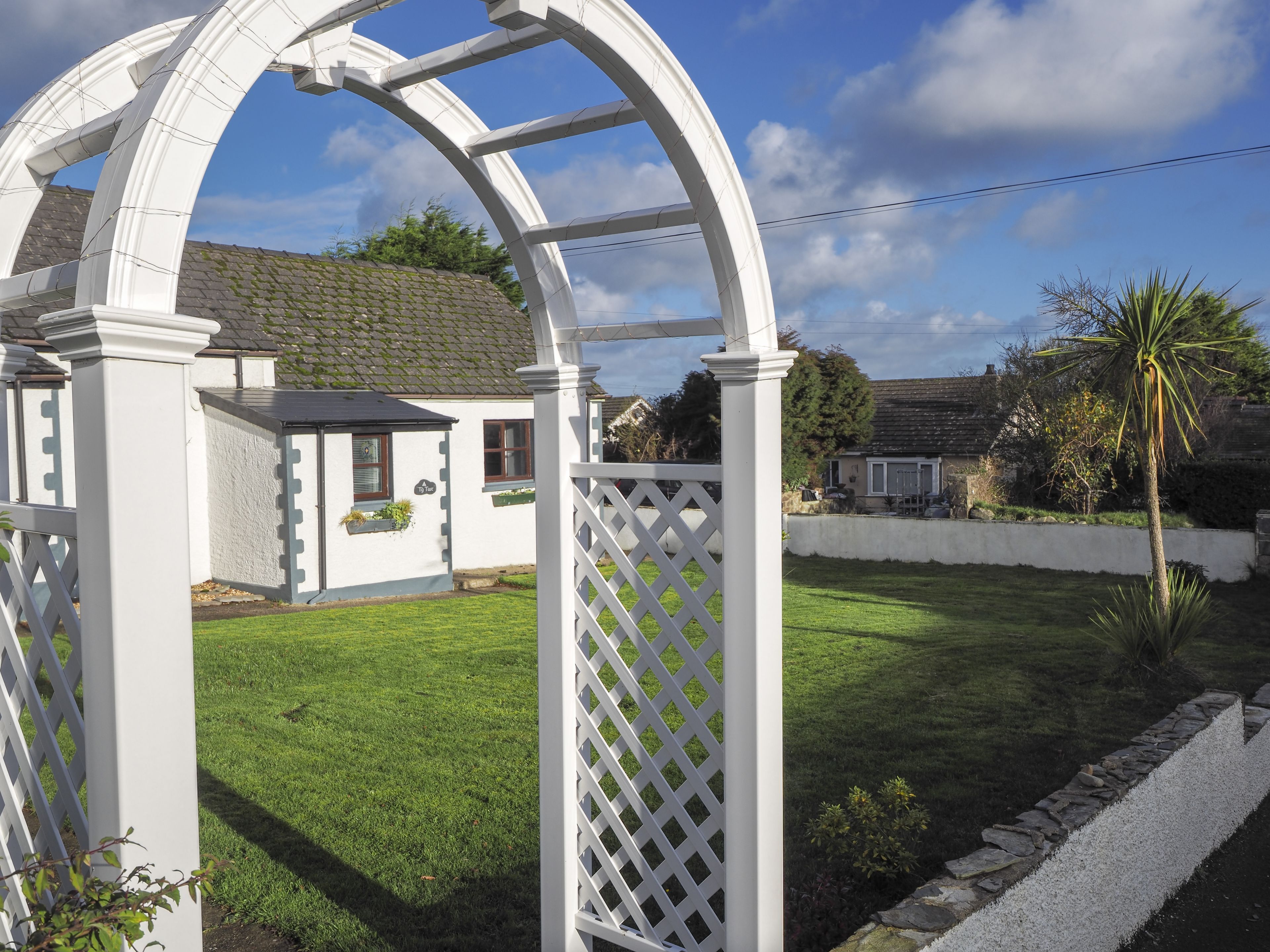 Een witte houten pergola in een tuin, met een huis op de achtergrond.