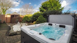 A hot tub in a backyard with a wooden deck, a stone fountain, and various shrubs and trees.
