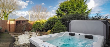 A hot tub in a backyard with a wooden deck, a stone fountain, and various shrubs and trees.