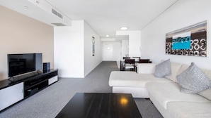 A modern living room with a flat-screen TV, a dark coffee table, and a white sofa with patterned cushions. - Sierra Broadbeach - West Hinterland View (Broadbeach)