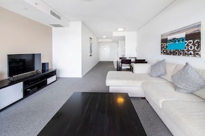 Ein modernes Wohnzimmer mit Fernseher, einem dunklen Couchtisch und einem weißen Sofa mit gemusterten Kissen.