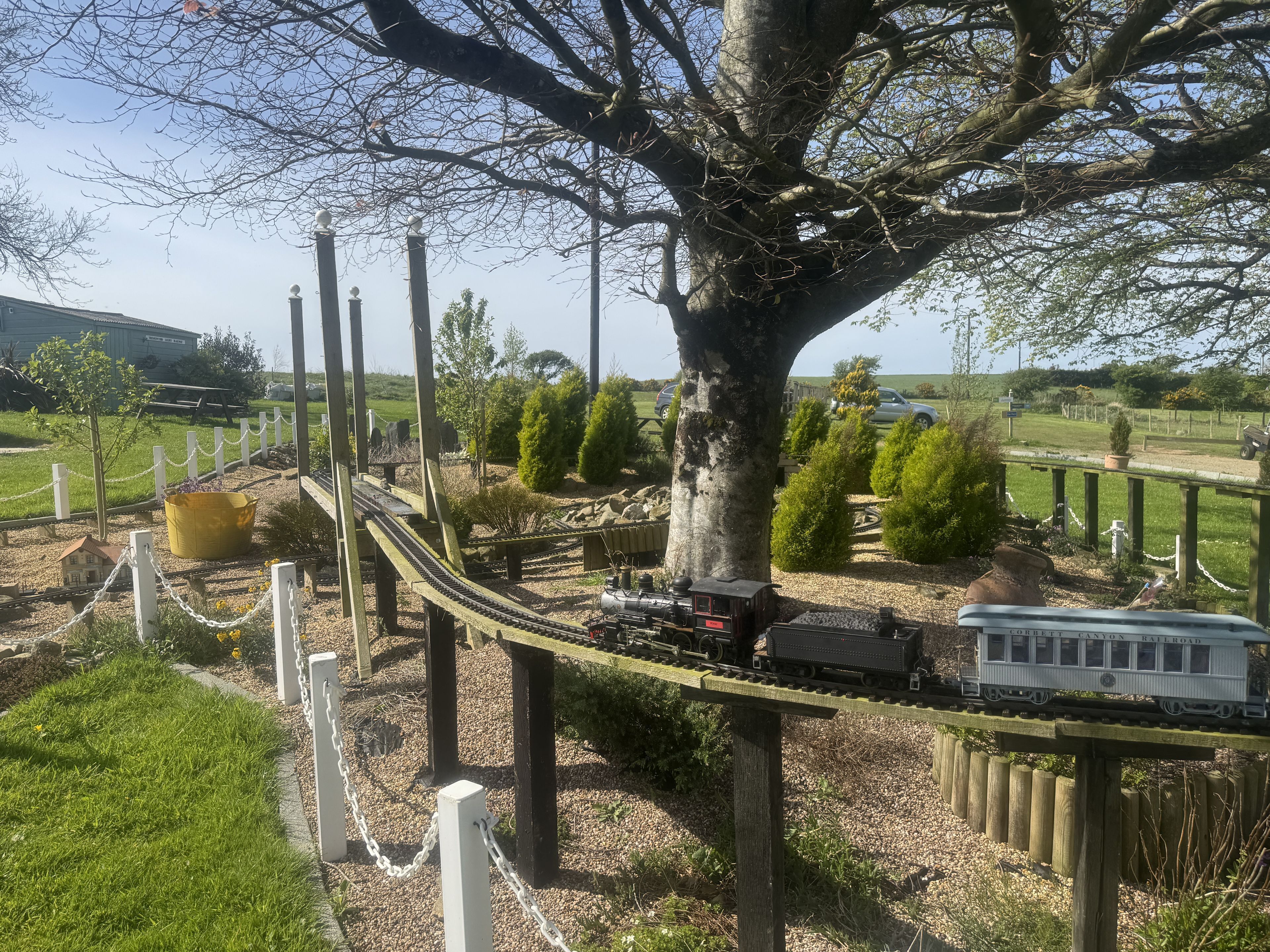 A model train set with a tree and a small house, surrounded by a fence and greenery.
