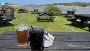 Een picknicktafel met twee drankjes, een helder glas bier en een donkere frisdrank met rietje. De tafel staat buiten, met uitzicht op een strand en de zee op de achtergrond.