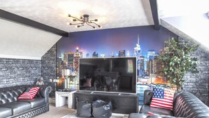 Een woonkamer met een grote flatscreen televisie, zwarte leren banken met kussens met de Amerikaanse vlag, een kleine salontafel, en een muurschildering van een stadsgezicht.