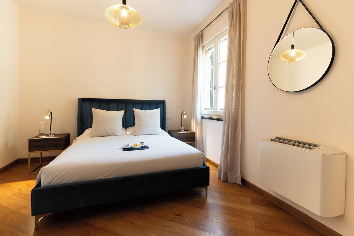 A neatly made bed with pillows, a bedside table with a lamp, a wall-mounted mirror, and a window with curtains.