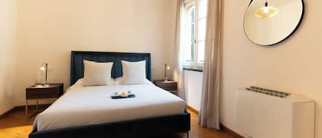 Un letto rifatto con cuscini, un comodino con una lampada, uno specchio fissato alla parete e una finestra con tende.