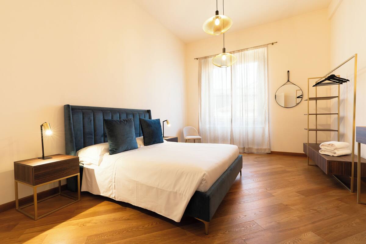 A bedroom with a bed, bedside tables, a hanging light fixture, a mirror, a wardrobe, and a window with curtains.