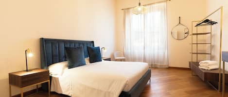 Quarto com cama, mesas de cabeceira, luminária pendente, espelho, guarda-roupa e janela com cortinas.