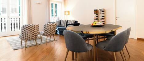 A dining area with a round table, four chairs, a bookshelf, and a balcony with curtains.
