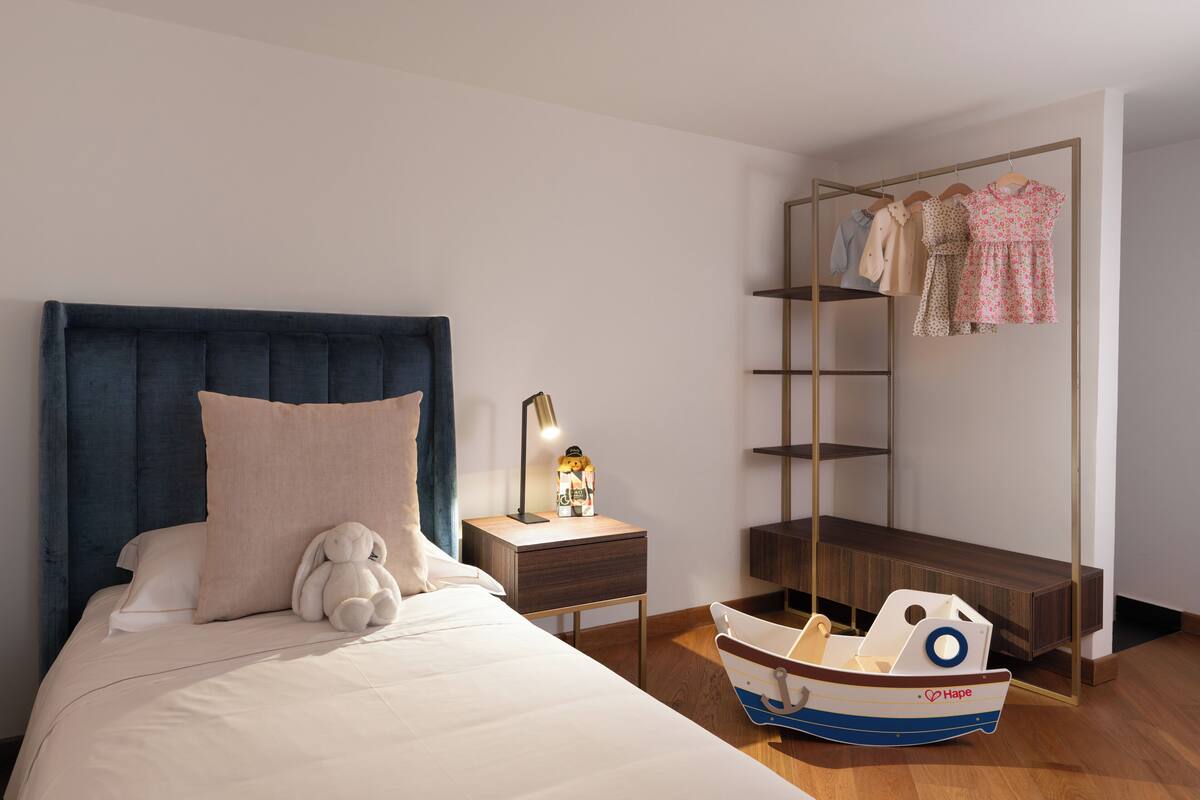 A bedroom with a bed, a nightstand, a lamp, a toy boat, and a clothing rack.