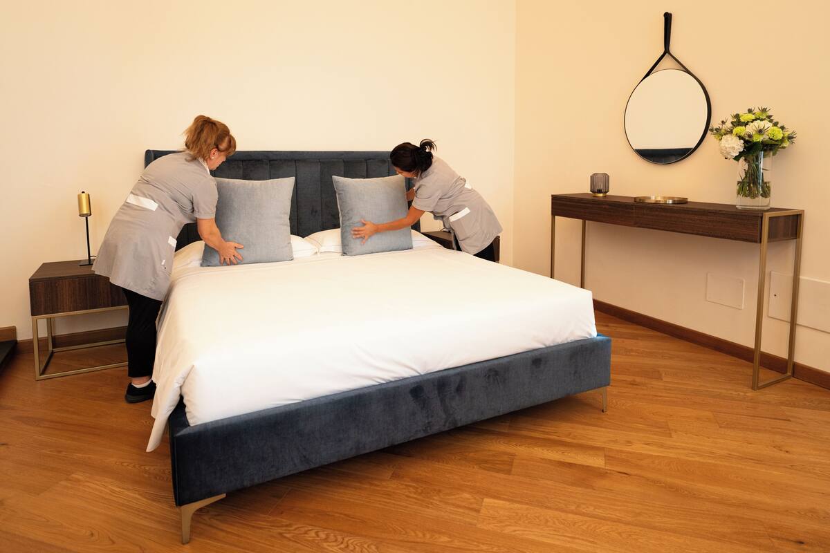 A neatly made bed with a dark headboard, a bedside table with a lamp, and a vase with flowers on a console table.